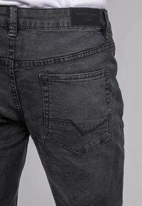 Jean en denim noir avec une poche arrière ornée d'un motif en chevron, agrémenté de détails de couture subtils et d'une étiquette en cuir au niveau de la taille.