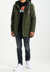 Olivově zelená vrstvená bunda s kapucí, předními kapsami a bílými šňůrkami, nošená přes černé tričko s potiskem a tmavé džíny, doplněná bílými botami.