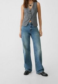 Szara kamizelka bez rękawów z zapięciem na guziki, w zestawie z wysokim stanem i szerokimi nogawkami niebieskich dżinsów. Noszona z czarnymi butami wsuwanymi.