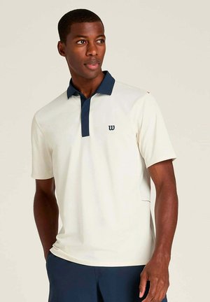 Mann trägt weißes kurzärmeliges Poloshirt mit dunkelblauem Kragen und marineblauer Hose, steht vor hellem einfarbigem Hintergrund und schaut nach rechts.