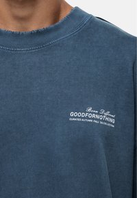 Sweatshirt en coton bleu avec un col rond, présentant des bords effilochés et un texte logo brodé blanc sur la poitrine.