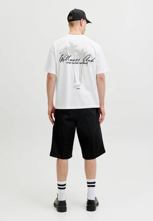 Mężczyzna noszący białą koszulkę z grafiką palm i tekstem, czarne szorty, czapkę, paskowane skarpetki i czarne buty, stoi tyłem.