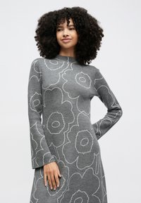 Žena s kudrnatými vlasy ve šedých šatech s velkými obrysy květinových motivů, stojící s jednou rukou na boku před jednoduchým pozadím.