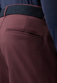 Pantalon bordeaux avec une texture lisse, doté d'une poche arrière avec une couture horizontale, et d'une ceinture noire texturée avec un détail de logo.