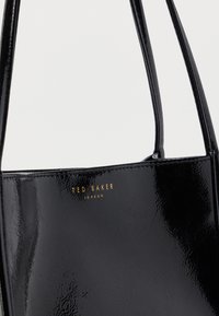 Sac fourre-tout en cuir verni noir avec deux poignées, arborant un logo doré "Ted Baker London" sur le devant et une texture lisse et brillante.