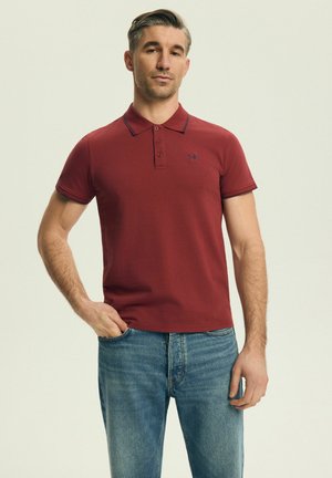 Mann trägt ein rotes kurzärmeliges Poloshirt und blaue Jeans, steht mit einer Hand in der Tasche vor neutralem Hintergrund.