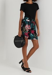 T-shirt noire associée à une jupe à imprimé floral avec des motifs roses et verts. Accessoires comprenant un sac rond noir et des sandales à talons.