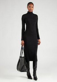 Zwart turtleneck jurkje met lange mouwen, aansluitende silhouet, zachte stof, gecombineerd met een glanzende zwarte handtas en kniehoge puntige laarzen.