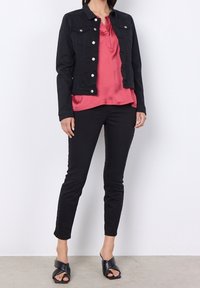 Chaqueta de mezclilla negra con botones metálicos, llevada sobre una blusa rosa. Combinada con pantalones negros ajustados y sandalias negras de tiras.