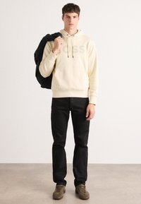 Sudadera con capucha beige con el logo "BOSS", pantalones negros y zapatillas verde oliva. El modelo sostiene una chaqueta negra sobre el hombro, de pie sobre un fondo neutral.