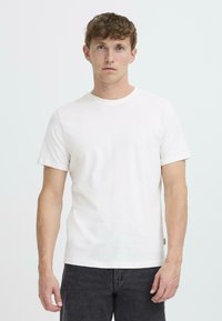 Camiseta de algodón blanca con cuello redondo y mangas cortas, que presenta un pequeño detalle de logo en el pecho. Combinada con jeans de color gris oscuro.