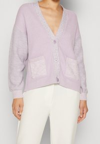 Ljust lila cardigan med V-hals, två framfickor och ribbad textur på kroppen. Har släta ärmar och knappstängning.