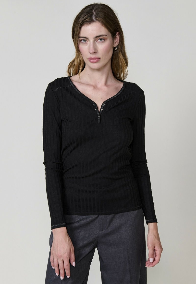 Haut noir à manches longues avec une texture côtelée, présentant un décolleté en V et un plastron à boutons à l'avant, associé à un pantalon gris.