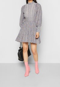 Žena v květinových šatech s dlouhým rukávem a krajkovými detaily, držící černou kabelku a oblečená v růžových ponožkových bot�ách, stojí proti jednobarevnému pozadí.