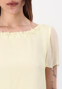 Blouse jaune à manches courtes et transparentes, avec un col en appliqué floral. Fabriquée en tissu texturé, elle a une coupe ample et une belle fluidité.