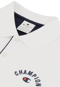 Polo blanca hecha de tela texturizada. Cuenta con un cuello azul marino y un logo bordado en azul marino y naranja en el pecho. Tiene una tapeta de dos botones.
