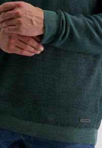 Hände sind über einen dunkelgrünen gestrickten Pullover mit gerippten Bündchen und Saum gefaltet, der mit blauen Jeans getragen wird.