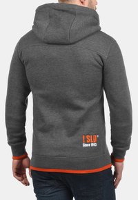 Sweat à capuche gris avec des accents orange, arborant un logo au dos. Le matériau semble doux avec une coupe classique et une capuche réglable.