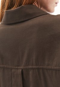 Camicia marrone con una texture morbida, dotata di un colletto classico e un dettaglio cucito sul retro. Il tessuto appare leggero e liscio.