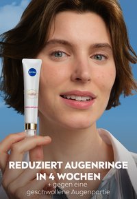 NIVEA eye care product in a white tube with gold accents, featuring “Luminous” label. Model with short brown hair holds it up against a blue background.