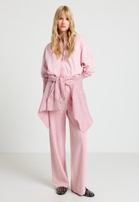 Junge Frau in einem hellrosa Trainingsanzug mit einem gestreiften Hemd, das um die Taille gebunden ist, und schwarzen, mit Nieten besetzten Schuhen, stehend vor einem schlichten weißen Hintergrund.