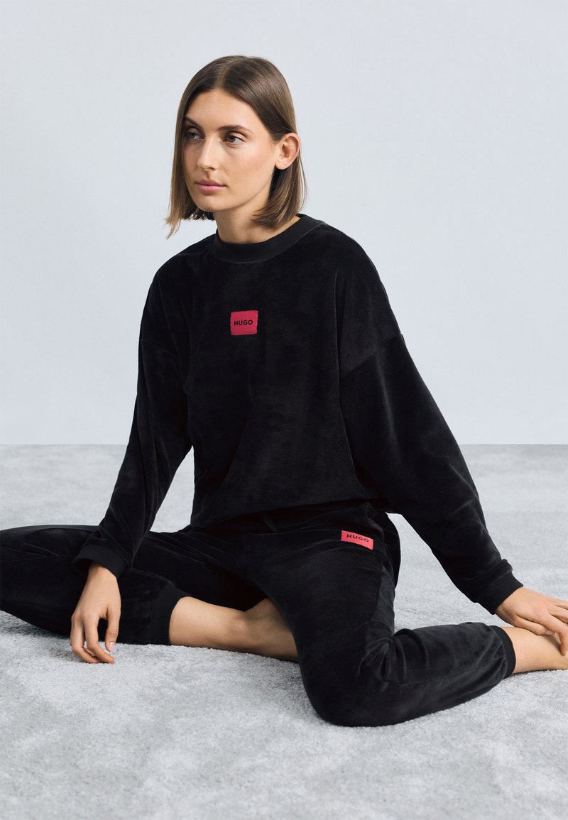 Čierny velúrový loungewear set pozostávajúci z dlhého rukávu a nohavíc s červeným štvorcovým logom "HUGO". Hladká textura, voľný strih.