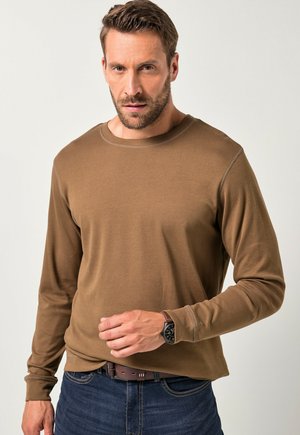 Braunes Langarmshirt aus weichem Material mit rundem Ausschnitt, glatter Textur und kontrastierenden Nähten. Getragen mit blauen Jeans und einer Uhr.