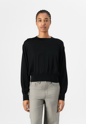 Svart langermet topp med rund hals og elastisk kant. Myk stoff; tettsittende i midjen, løse ermer. Sammen med lys grå bukser.