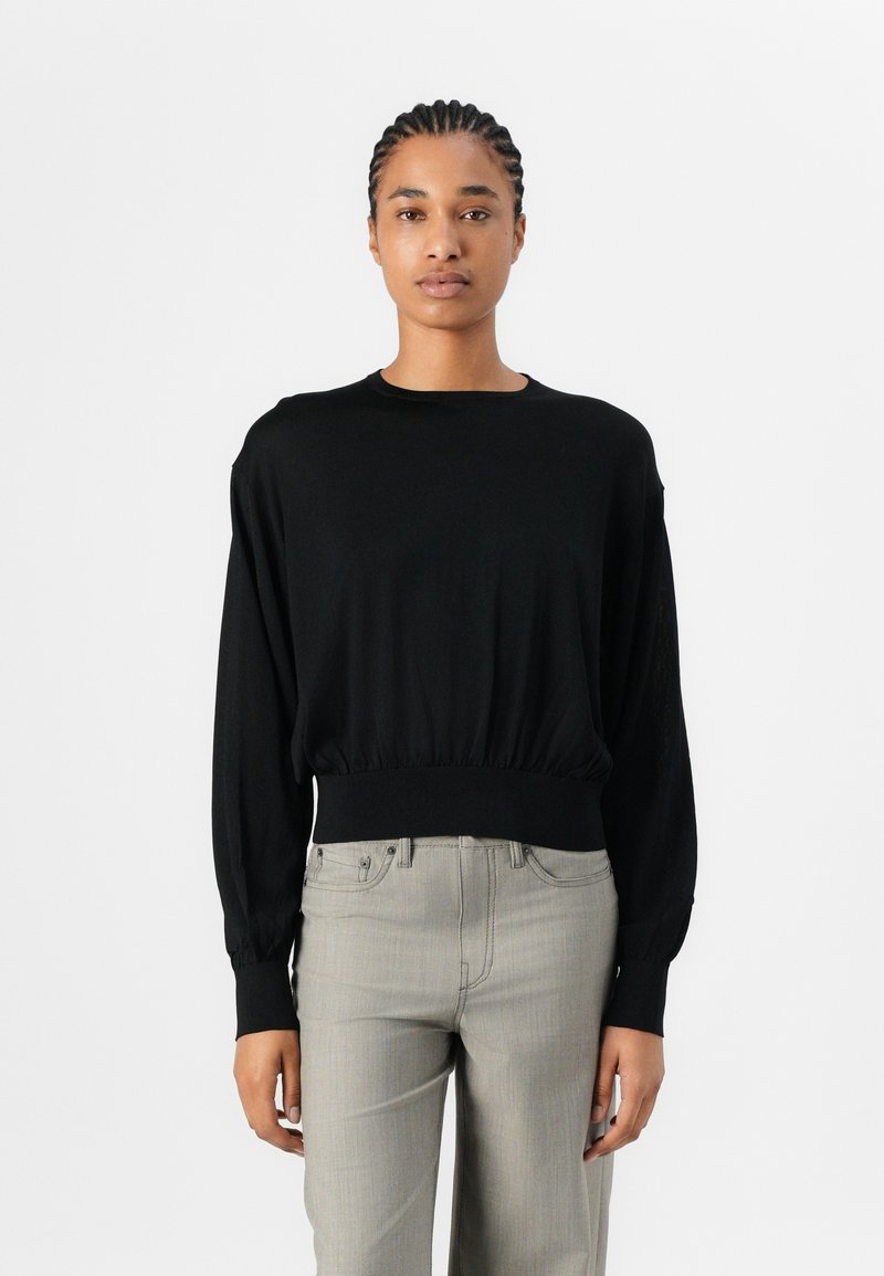 Black long-sleeve top with a round neckline and elasticized hem. Soft fabric; fitted at the waist, loose-fitting sleeves. Paired with light grey pants.