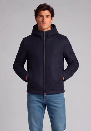 Jonge man met een zwarte zip-up hoodie, handen in de zakken en blauwe spijkerbroek, staand tegen een effen lichtgrijze achtergrond.