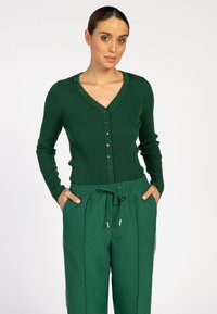 Groene geribde cardi met een V-hals, voorzien van knoopdetails langs de halslijn. Gecombineerd met bijpassende groene broek met koordjes.