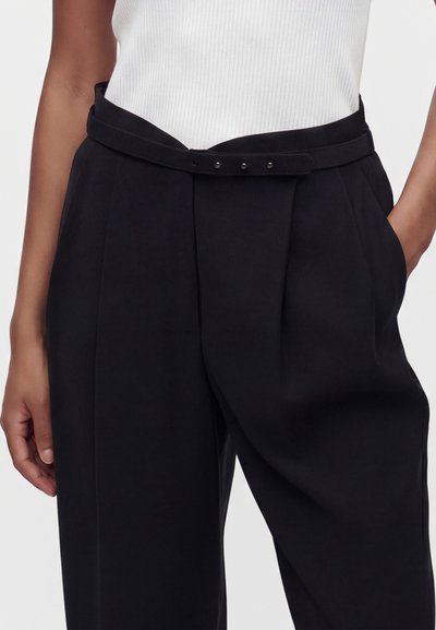 Άτομο που φορά μαύρο παντελόνι με πιέτες και ψηλή μέση, ασορτί ζώνη και λευκή ριγέ αμάνικη μπλούζα, με το ένα χέρι στην τσέπη.