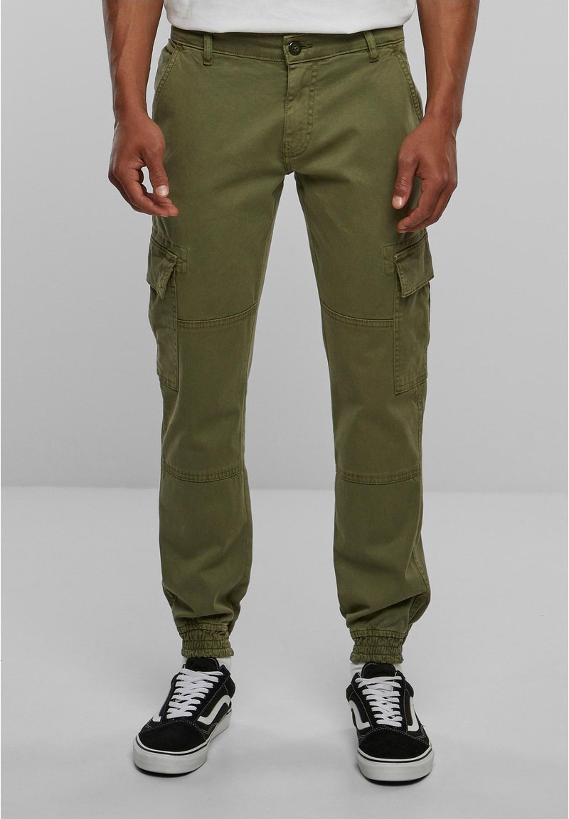 Olijfgroene cargo broek met elastische boorden, voorzien van meerdere zijzakken en een zachte textuur. Gecombineerd met zwarte sneakers en witte veters.