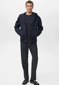 Sudadera con capucha azul marino y cremallera, con bolsillos tipo canguro, combinada con una camiseta ajustada de cuello redondo negra y pantalones grises a medida. Zapatos negros completan el conjunto.