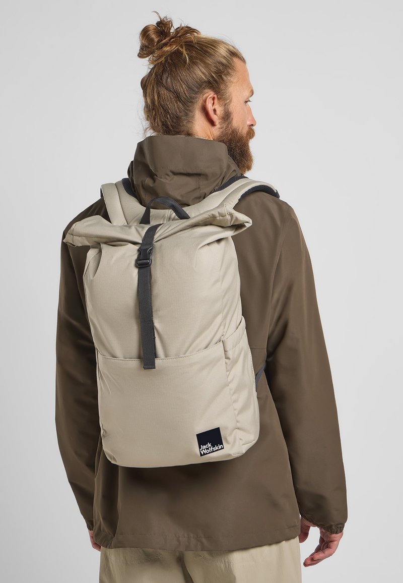 Beige rugzak met een oprolbaar ontwerp, verstelbare banden en een voorzak. Gemaakt van duurzaam fabric met een klein logo-label aan de onderkant.