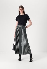 Fustă plisată metalică de culoare verde închis, purtată cu un tricou negru, completată de sandale masive negre și o geantă mică de umăr neagră.
