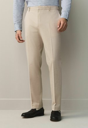 Man wearing beige dress trousers, light blue patterned shirt, and black leather dress shoes, standing against a plain light wall.