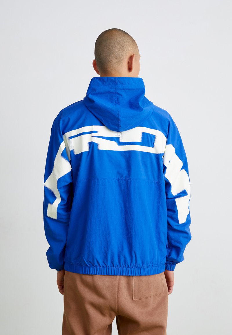 Kék, könnyű dzseki kapucnival és fehér geometrikus mintával a hátsó részen. Rugalmas alsó szegély és mandzsetta, sima textúrájú.