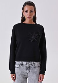 Felpa nera con scollatura tonda, caratterizzata da applicazioni floreali nere in rilievo sul davanti e polsini a coste. Abbinata a jeans di denim chiaro.