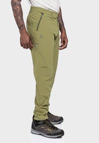 Olivgrüne Wanderhosen mit schlanker Passform, Reißverschluss-Taschen und verstärkten Nähten. Kombiniert mit schwarzen Wanderschuhen mit gelben Akzenten.