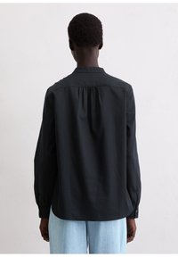 Czarna koszula z długim rękawem, zapięty kołnierzyk na guziki, marszczony tył i lekko luźny krój; rękawy zakończone mankietami na guziki. Miękki, gładki materiał.