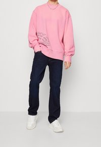 Sudadera rosa oversize con un diseño texturizado en gris en la parte delantera, combinada con jeans oscuros y sneakers blancos. Corte relajado con puños acanalados.