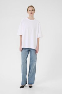 Tricou alb oversized din bumbac, cu mâneci scurte, asortat cu blugi drepți de color bleumarin deschis și pantofi cu vârf ascuțit negri. Fundal simplu.