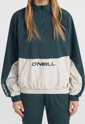 Personne portant un pullover polaire O’Neill vert et beige avec zip quart et poche avant, debout les mains relâchées le long du corps.