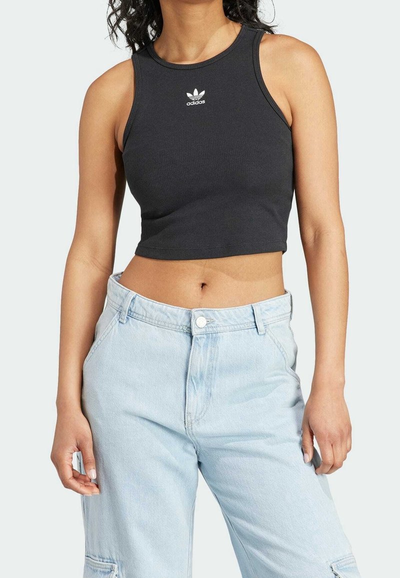 Kvinna i en svart Adidas croppad tanktop och ljusblå högmidjade jeans som står med avspända händer.