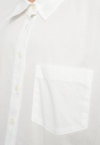 Bela srajca s knappami, izdelana iz lahkotnega blaga. Ima ovratnik z ostrimi konci, šest gumbov in žep na levi strani prsi z ravnim robom.