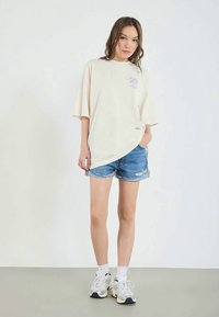 T-shirt beige oversize con grafica di palme viola, abbinato a pantaloni corti in denim strappati. Il modello indossa calzini bianchi e sneaker chunky.