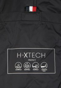 Étiquette H-XTECH sur tissu noir indiquant les caractéristiques imperméables, respirantes, coupe-vent, et icônes des coutures principales étanches.