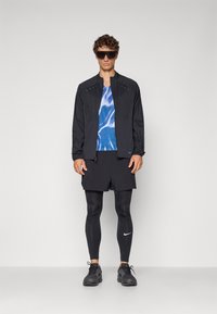 Schwarze leichte Jacke mit Reißverschluss, blau gemustertes Hemd, schwarze Shorts und Kompressionsleggings, kombiniert mit schwarzen Turnschuhen und Sonnenbrille.