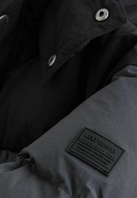 Black and gray padded jacket featuring a textured fabric, snap buttons, and a rectangular brand label on the sleeve.